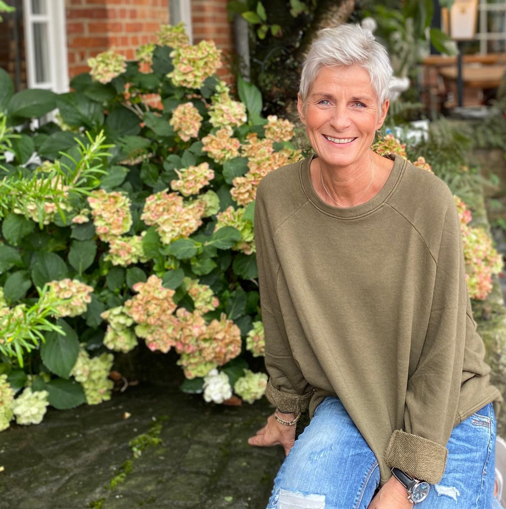 Foto von einer Frau mit blonden Haaren, Petra Battmer im grünen Pullover mit Jeans vor gelben Blumen